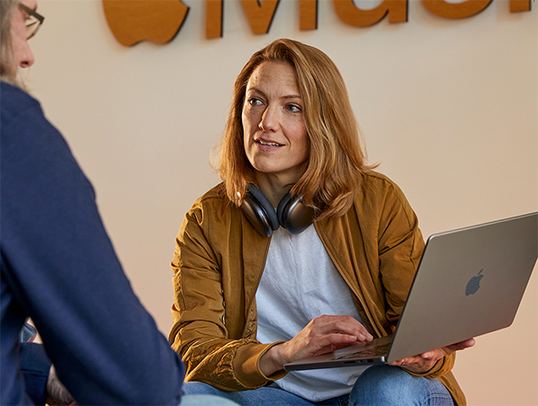 Two Apple Sales and Business Development team members in conversation, one holding a MacBook and sitting in front of a wall with the Apple Music logo on it.