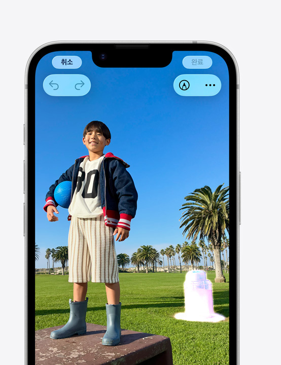 공원의 낮은 돌담 위에 서서 웃고 있는 사람의 모습이 담긴 사진에서 클린업 도구로 배경의 쓰레기통을 지우는 방법을 보여주는 iPhone 화면