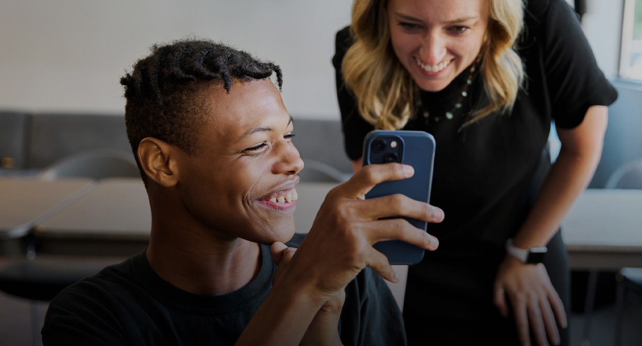 Photo d’un jeune homme malvoyant qui sourit en montrant l’écran de son iPhone à une femme souriante qui se tient derrière son épaule gauche