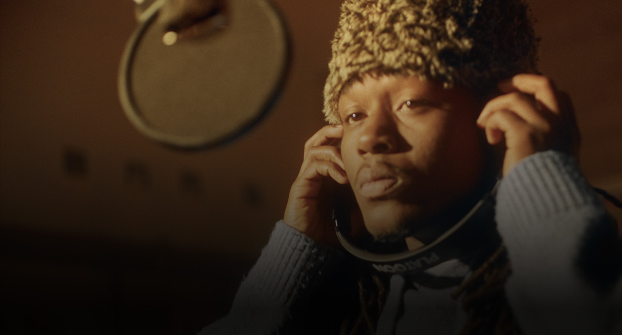 Photo de Kiddo K dans un studio de musique debout devant un micro, écoutant la musique dans un casque