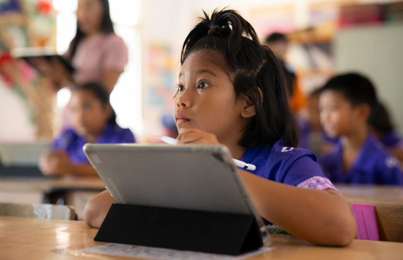A young girl sits at her desk, looking towards the front of the classroom and fully engaged. She is using an iPad and holding an Apple Pencil.
