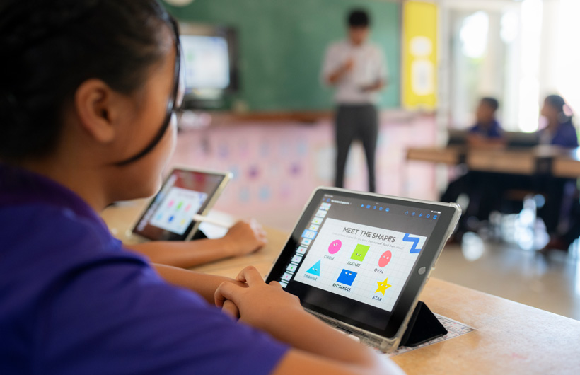 Close-up of a student at their desk, using an iPad. On screen, there are colourful shapes displayed with the title, “Meet the shapes.”