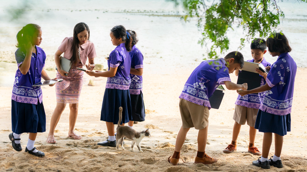 Six primary school students in colourful purple uniforms stand on a sandy beach holding iPads. A teacher is assisting one student on an iPad, and there is a cat walking amongst them.