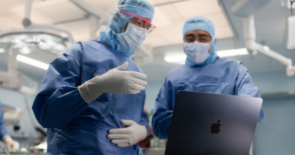 Two surgeons in the operating room look at a MacBook, and one gestures with their hand while the other observes