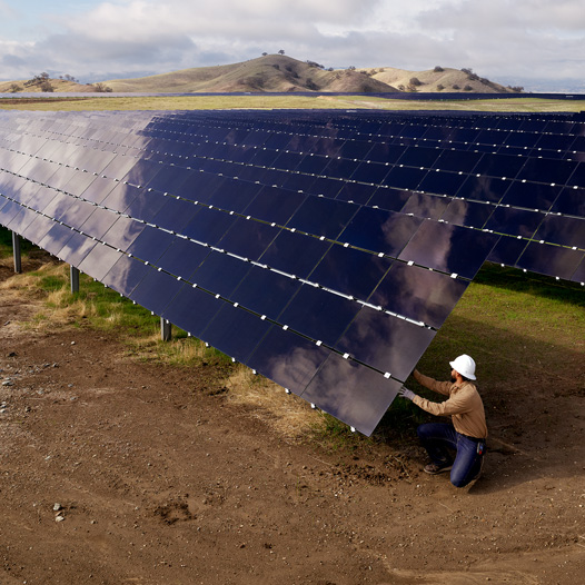 An employee in a safety hat inspects solar array that is in a field with hills in the near horizon